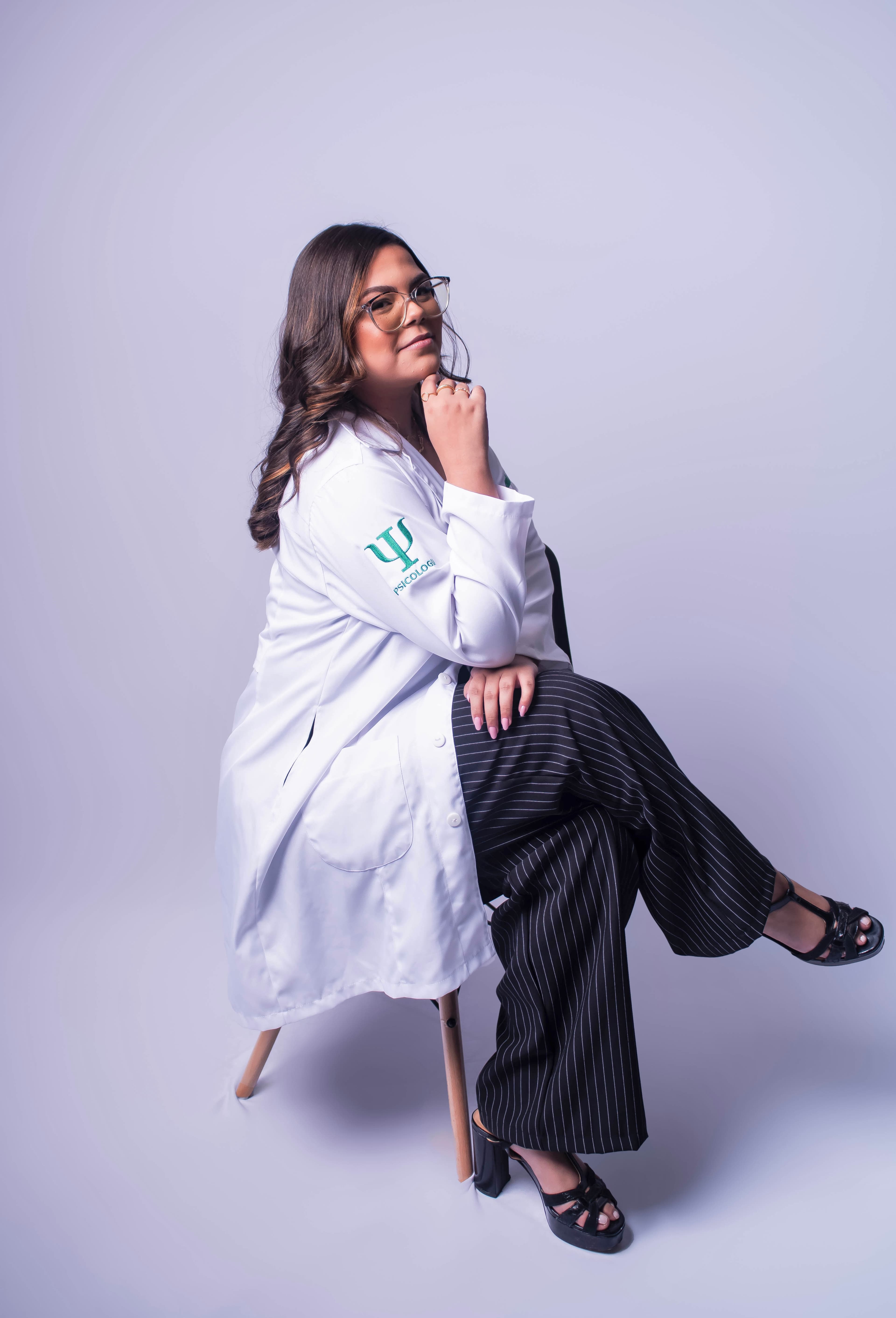 Maria Vitória de Souza do Rêgo, psicóloga, sentada em um banco de madeira, de jaleco branco com o símbolo da psicologia bordado em verde, apoiando o queixo na mão.